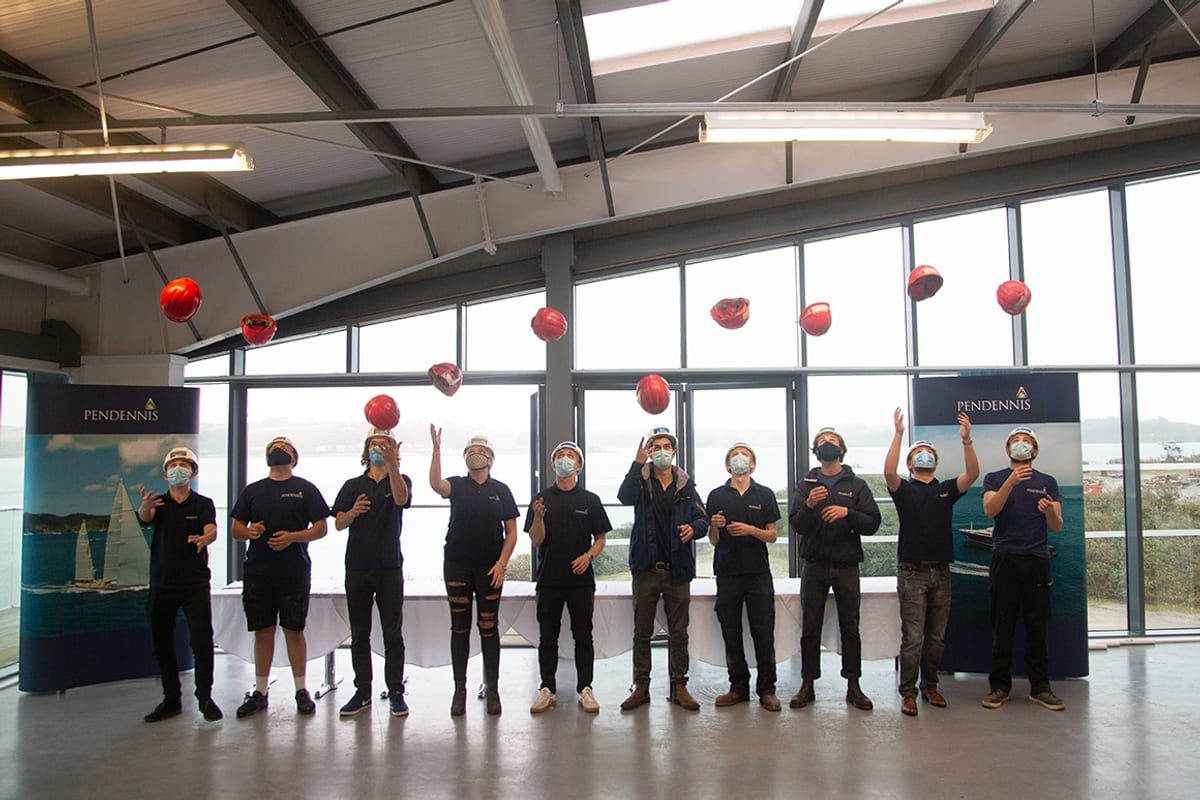 Pendennis apprentices wearing masks celebrate graduation indoors by tossing red hard hats into the air during the 2020 ceremony.