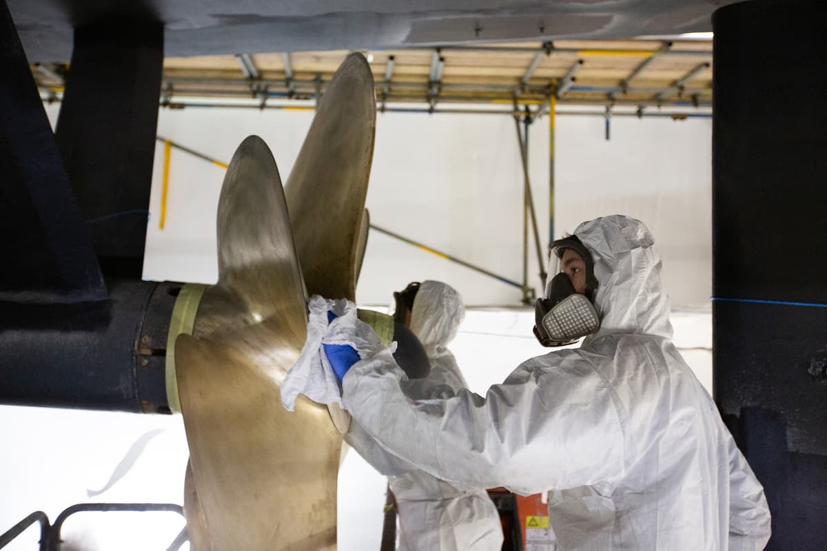 Pendennis Shipyard apprentice cleaning a superyacht propeller in preparation for Propspeed coating. Wearing full protective gear including a respirator and coveralls, he works with precision beneath the vessel in a dry dock.