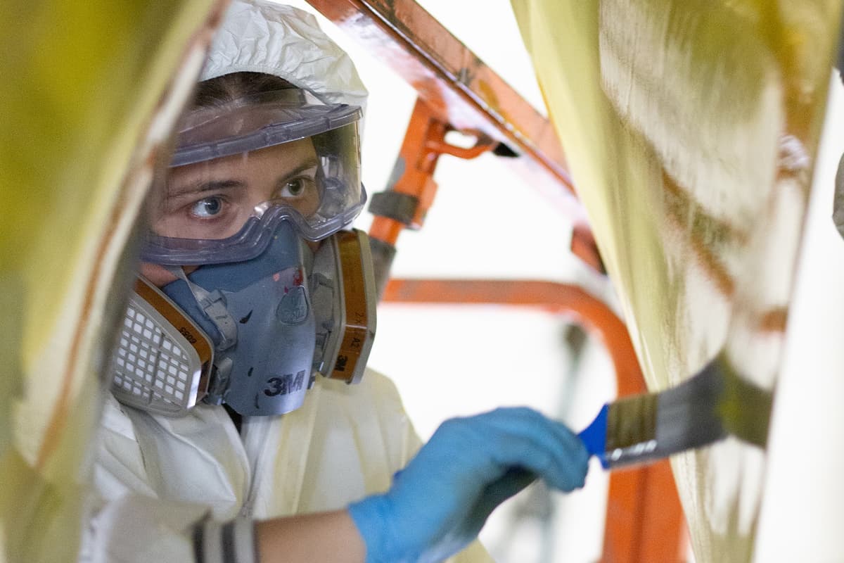 Pendennis Shipyard apprentice applying protective coating to a superyacht propeller. Wearing full PPE including a respirator and gloves, she carefully brushes on the coating while elevated on a platform.