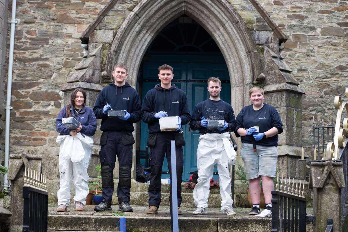 Pendennis Shipyard apprentices standing outside a historic stone building, holding paint trays and brushes. Wearing a mix of overalls and protective gloves, they pose in front of an arched doorway, ready for restoration or maintenance work.