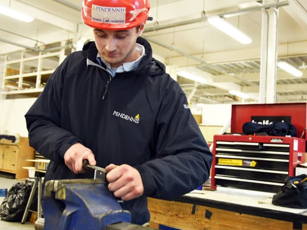 Pendennis Shipyard apprentice using a hand file on a piece of metal clamped in a workshop vice. Wearing a red safety helmet and branded jacket, he concentrates on refining the material in a well-equipped training space.