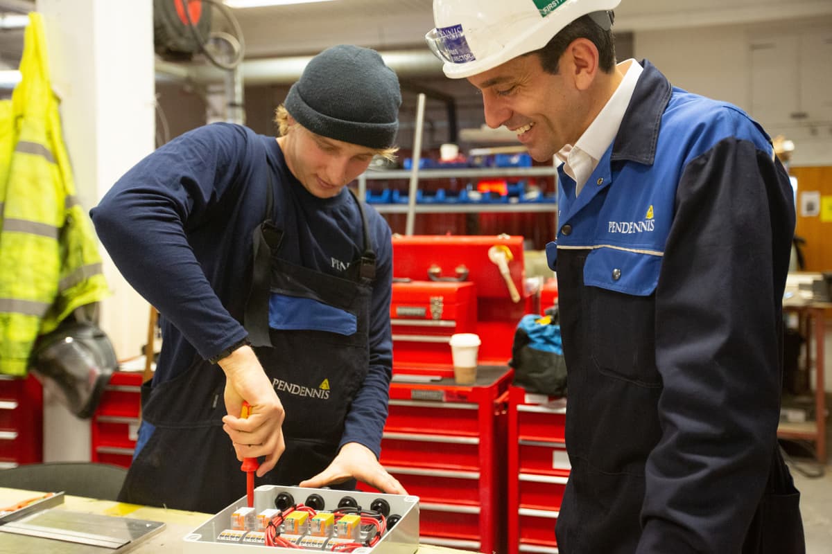 Pendennis Shipyard apprentice assembling an electrical unit while being observed by a senior team member. Both are smiling during the hands-on session in the workshop, surrounded by red tool chests and electrical components.