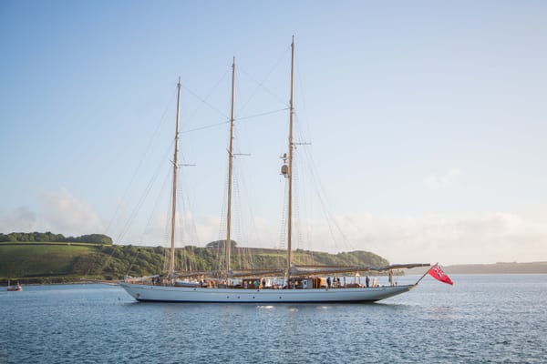 Three-masted sailing yacht Adix anchored in calm waters near the Cornish coast under clear evening light.