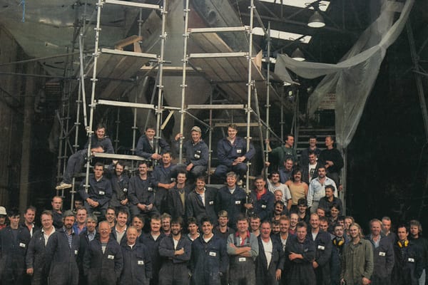 Pendennis Shipyard team pose beneath classic yacht Adela during her restoration, surrounded by scaffolding in the workshop.