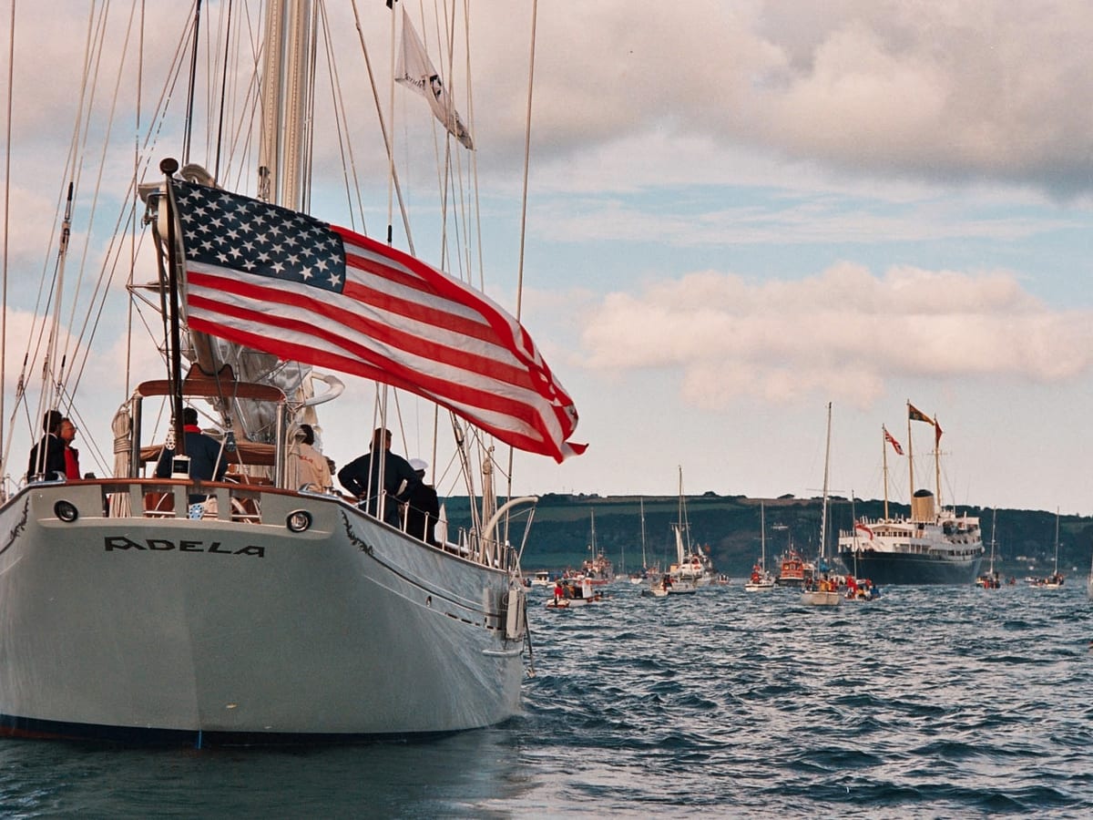 Classic sailing yacht Adela with American flag, followed by a fleet of boats and The Royal Yacht Britannia in the distance.