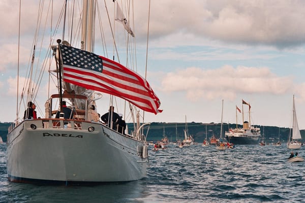 Classic sailing yacht Adela with American flag, followed by a fleet of boats and The Royal Yacht Britannia in the distance.