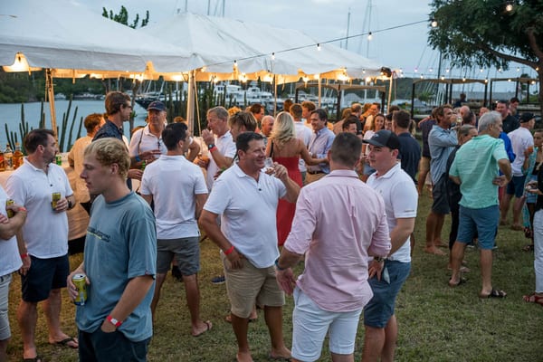 Guests gather under string lights and canopy tents at the waterfront welcome party, chatting, laughing, and enjoying drinks as the marina and superyachts provide a scenic Caribbean backdrop.