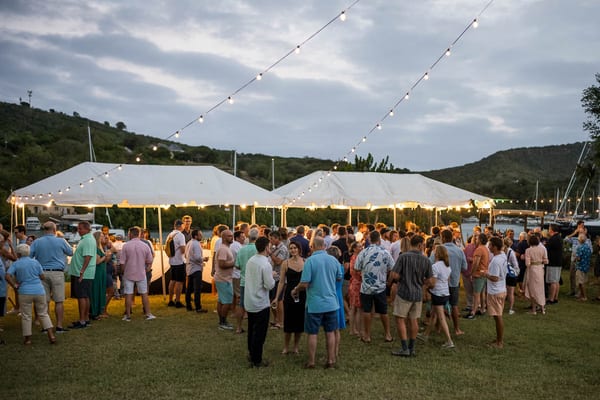 Guests gather under string lights and white canopy tents during an outdoor evening party by the water, with a relaxed crowd mingling and chatting as twilight settles over the hillside marina backdrop.