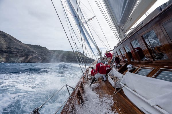 Crew members in matching red Adela shirts brace themselves on the polished wooden deck as the yacht heels over in heavy seas, with dramatic cliffs rising in the background.