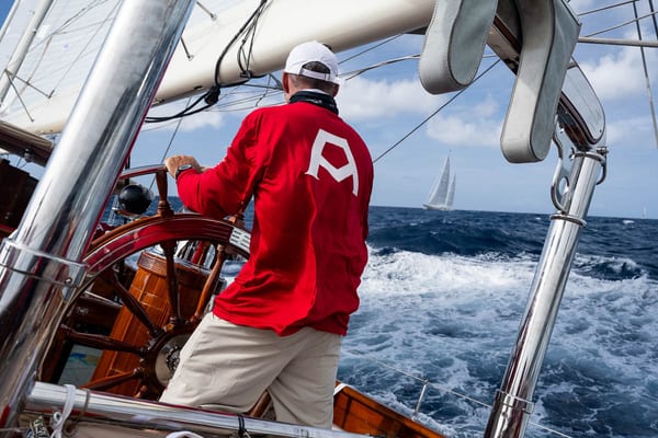 A crew member wearing Adela’s signature red kit helms the classic superyacht through rolling blue waves, one hand on her varnished wooden wheel, while another yacht sails in the distance under a bright Caribbean sky.