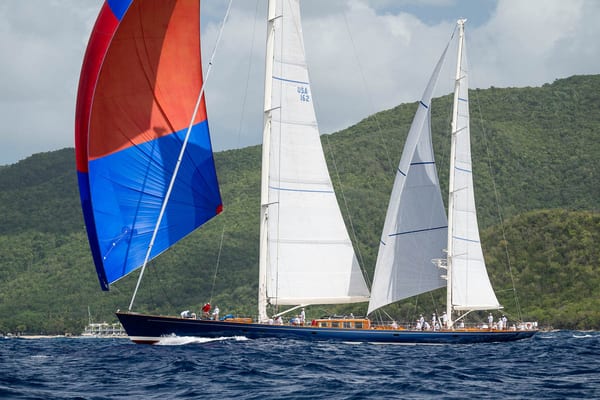 Superyacht Adela under sail with a striking blue and red spinnaker during the Antigua Superyacht Challenge 2025, cruising past the lush green coastline.