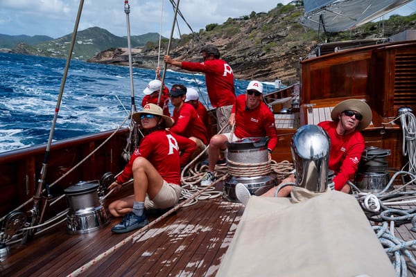 Crew of Adela in coordinated red shirts trimming sails during the Antigua Superyacht Challenge 2025, with rocky coastline in the background.