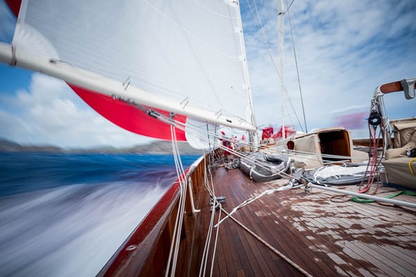 View from the deck of a classic sailing yacht at speed during the Antigua Superyacht Challenge 2025, with motion blur capturing the rush of wind and sea under red and white sails.