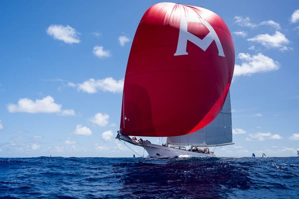 Classic superyacht Adela sails under a bright red spinnaker emblazoned with an 'A' during the Antigua Superyacht Challenge 2025, set against a vivid blue sky and ocean.