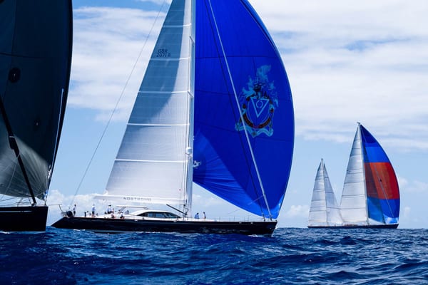 Superyacht Nostromo powers ahead under a blue spinnaker adorned with a crest, with Rebecca sailing behind during the Antigua Superyacht Challenge 2025.