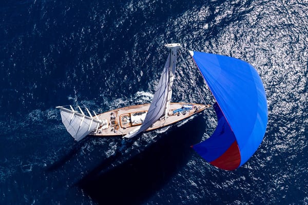 Aerial view of superyacht Rebecca racing under full sail with a blue and red spinnaker during the 2025 Antigua Superyacht Challenge, cutting a clear path through sparkling seas.