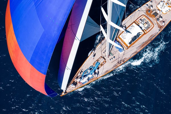 Aerial view of superyacht Rebecca under full sail, with a red and blue spinnaker billowing as crew work on the teak deck during the Antigua Superyacht Challenge.