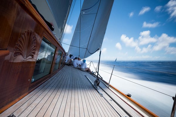 Crew sitting along the teak deck of superyacht Rebecca during a race, with sails full and motion blur of the sea showing speed.