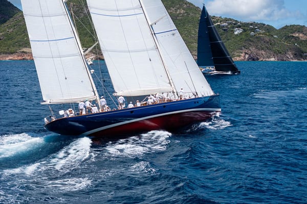 The classic ketch Rebecca sails close-hauled in turquoise waters during the 2025 Antigua Superyacht Challenge, with crew members on deck and another superyacht racing behind against the lush coastline.