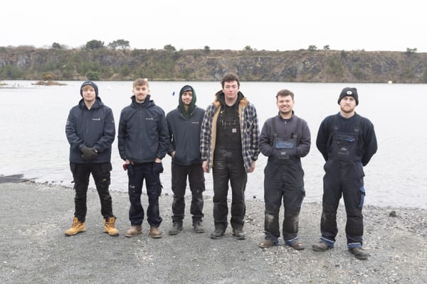 Group of Pendennis apprentices standing by the water at a quarry lake, dressed in outdoor workwear.