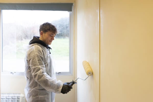 Pendennis apprentice painting an indoor wall with a roller while wearing disposable coveralls and gloves.