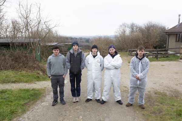 Five Pendennis apprentices standing outdoors in work gear and disposable coveralls, with countryside and picnic benches in the background.