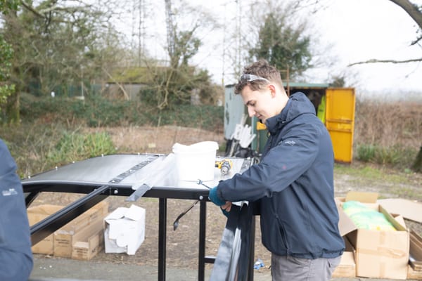 Pendennis apprentice assembling a curved metal frame outdoors, using hand tools beside open cardboard boxes and a drill.