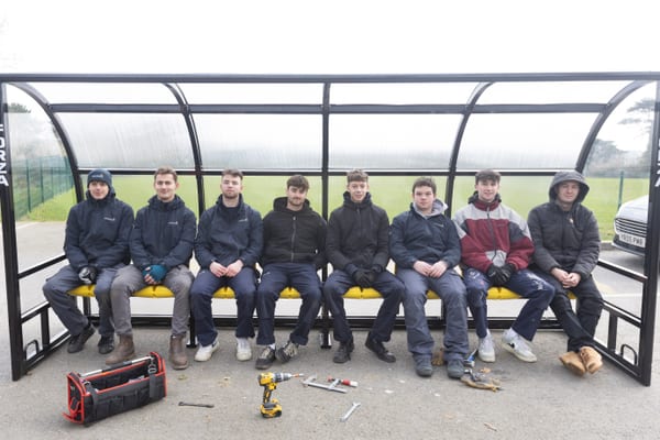 Eight Pendennis apprentices sitting side by side on a newly assembled dugout bench, with tools and a drill scattered on the ground in front.