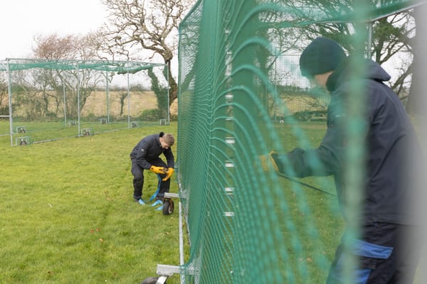 Two Pendennis apprentices securing netted fencing panels outdoors on a grassy field, wearing safety gloves and cold-weather gear.
