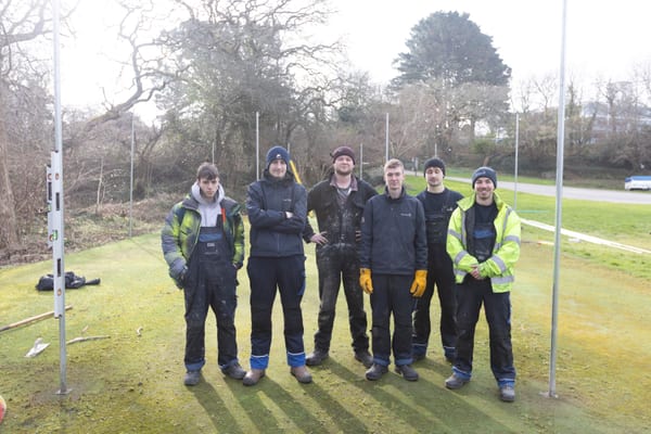 Group of Pendennis apprentices standing together outdoors after setting up tall metal posts on a grassy area.