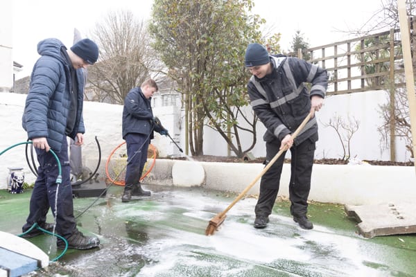 Pendennis apprentices cleaning an outdoor play area with a hose, pressure washer, and broom.