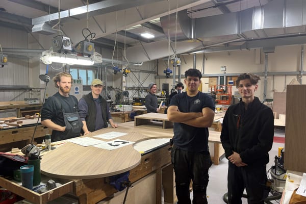 Pendennis apprentices and team members gathered around a curved woodwork bench in the joinery workshop.