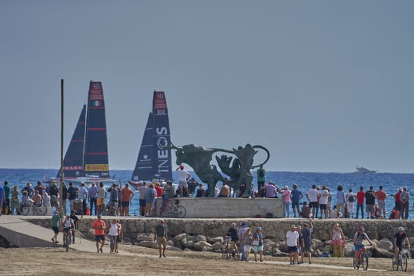Spectators watch America's Cup racing yachts sail past the El Pasifae sculpture on the Vilanova seafront.
