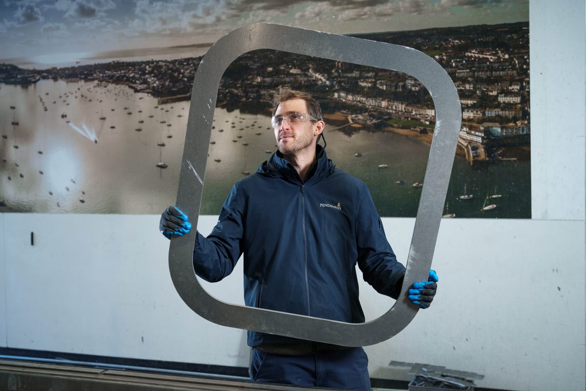 Technician holding a large metal component at Pendennis's Digital Manufacturing Centre in front of a Falmouth harbour mural.