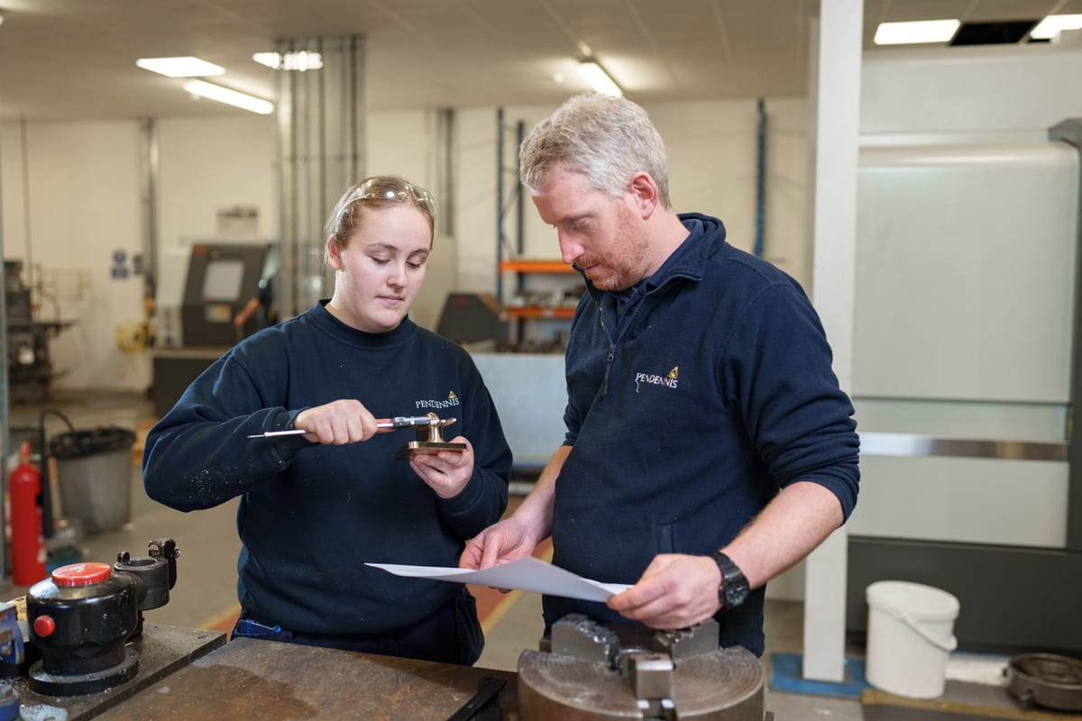 Apprentice using calipers to measure a machined part while being guided by an engineer at Pendennis.