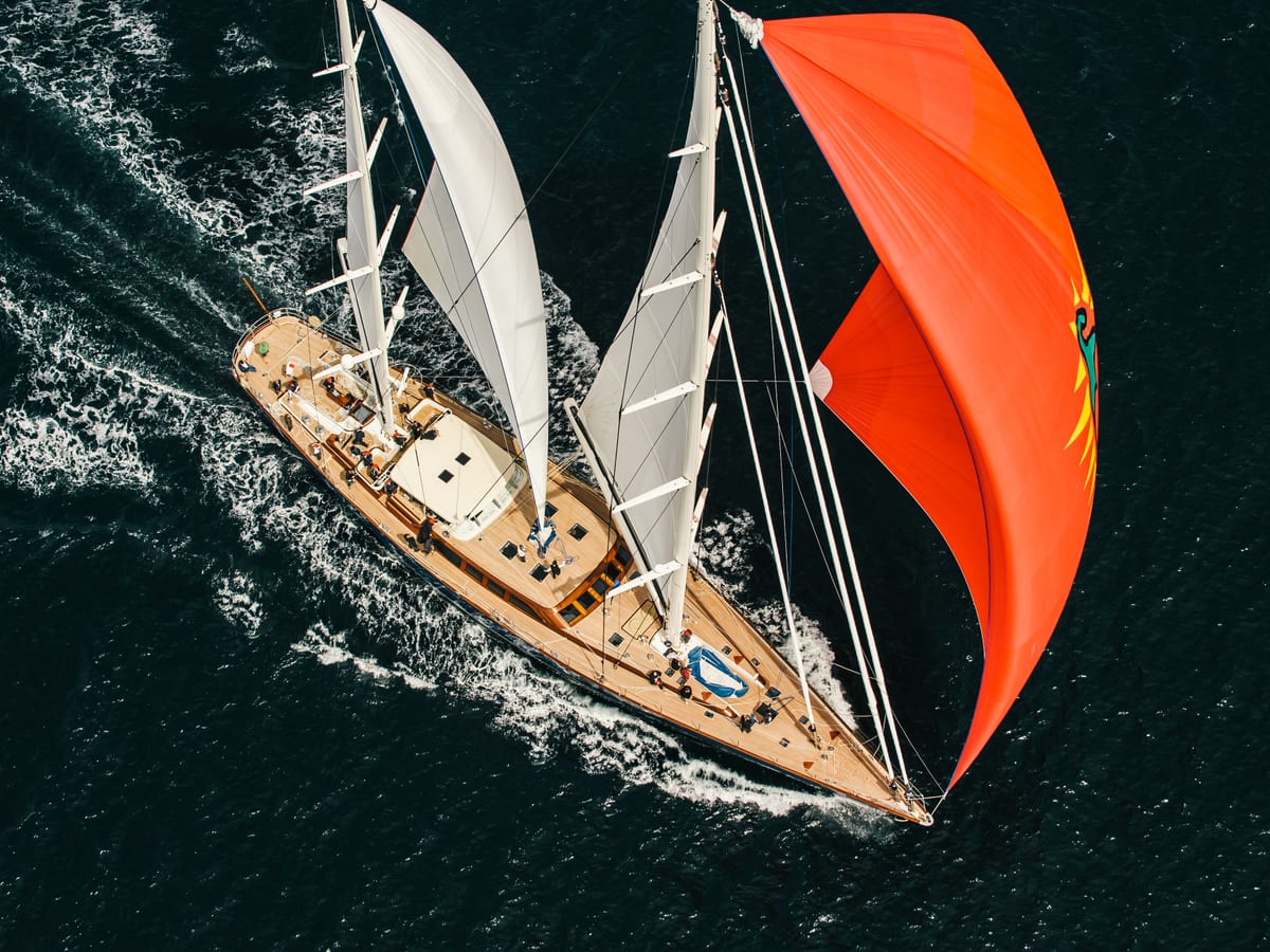Aerial view of classic sailing yacht Christopher with white sails and a striking red spinnaker cutting through deep blue water.