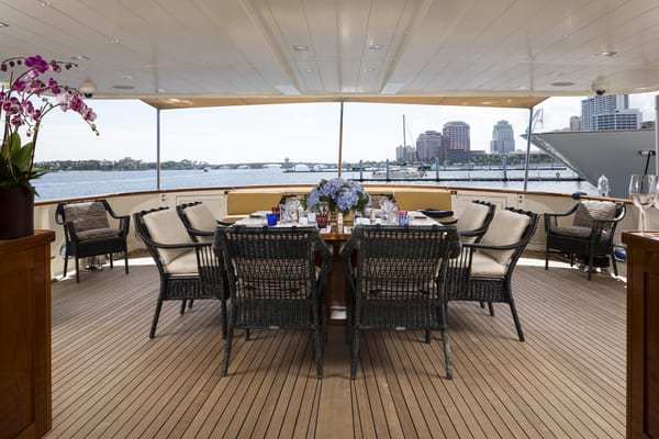 Aft deck dining area on motor yacht A2, featuring a set table with wicker chairs, waterfront views, and a city skyline in the background.