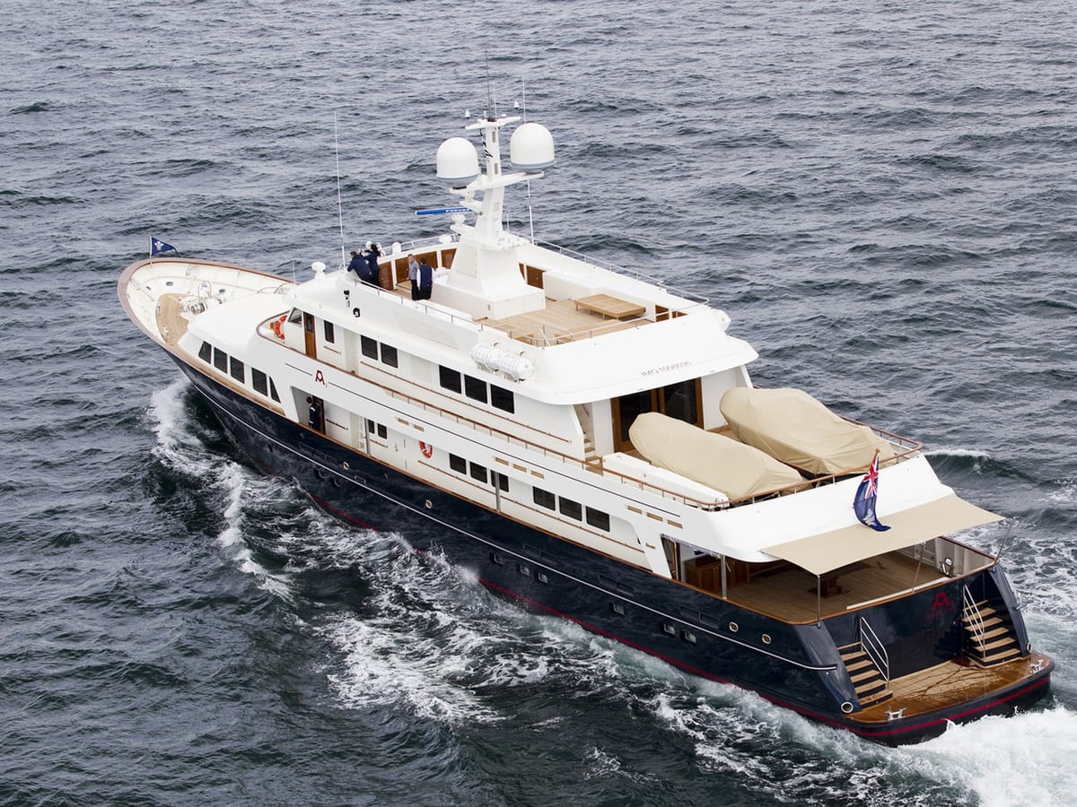 Aerial view of motor yacht A2 cruising at sea, showing her navy hull, white superstructure, covered tenders on the aft deck, and British ensign flag flying from the stern.