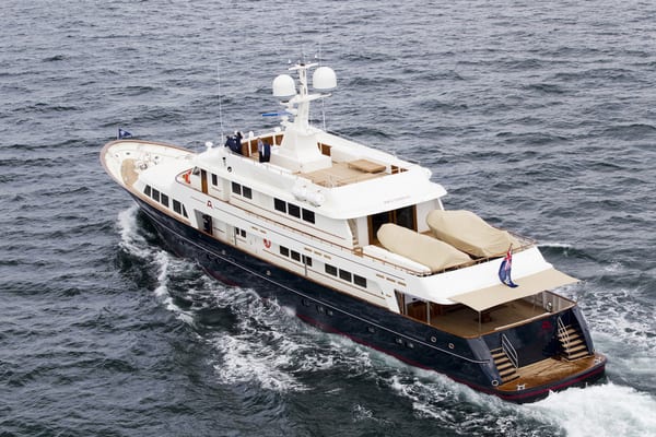 Aerial view of motor yacht A2 cruising at sea, showing her navy hull, white superstructure, covered tenders on the aft deck, and British ensign flag flying from the stern.