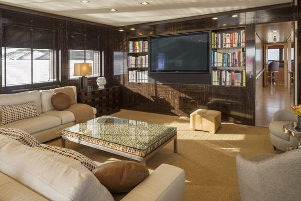 Luxurious yacht lounge with cream sofas, a glass coffee table, dark wood paneling, and built-in bookshelves surrounding a large flat-screen TV.