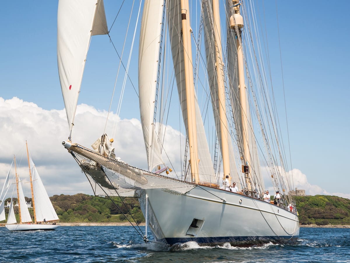 The classic schooner Adix sailing close to shore under full sail, with crew members on deck and another sailing yacht visible in the background near Falmouth.