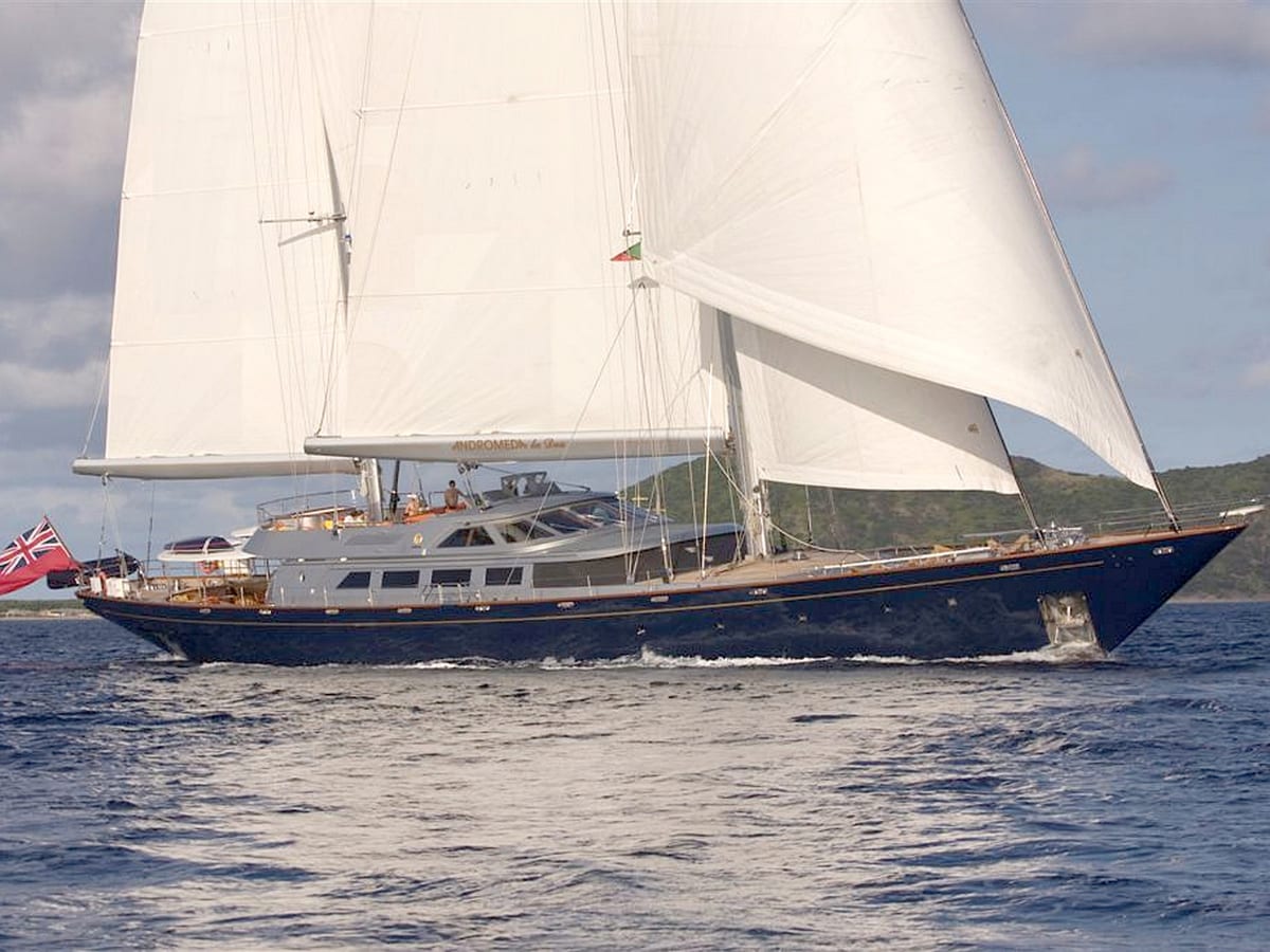 Sailing yacht Andromeda La Dea underway in the Caribbean, showing her sleek dark hull, white sails, and deck structures against a calm sea and green hills.