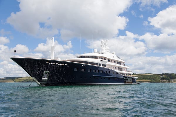 Aquila superyacht anchored off the coast near Falmouth, with her sleek black hull and white superstructure under a partly cloudy sky.