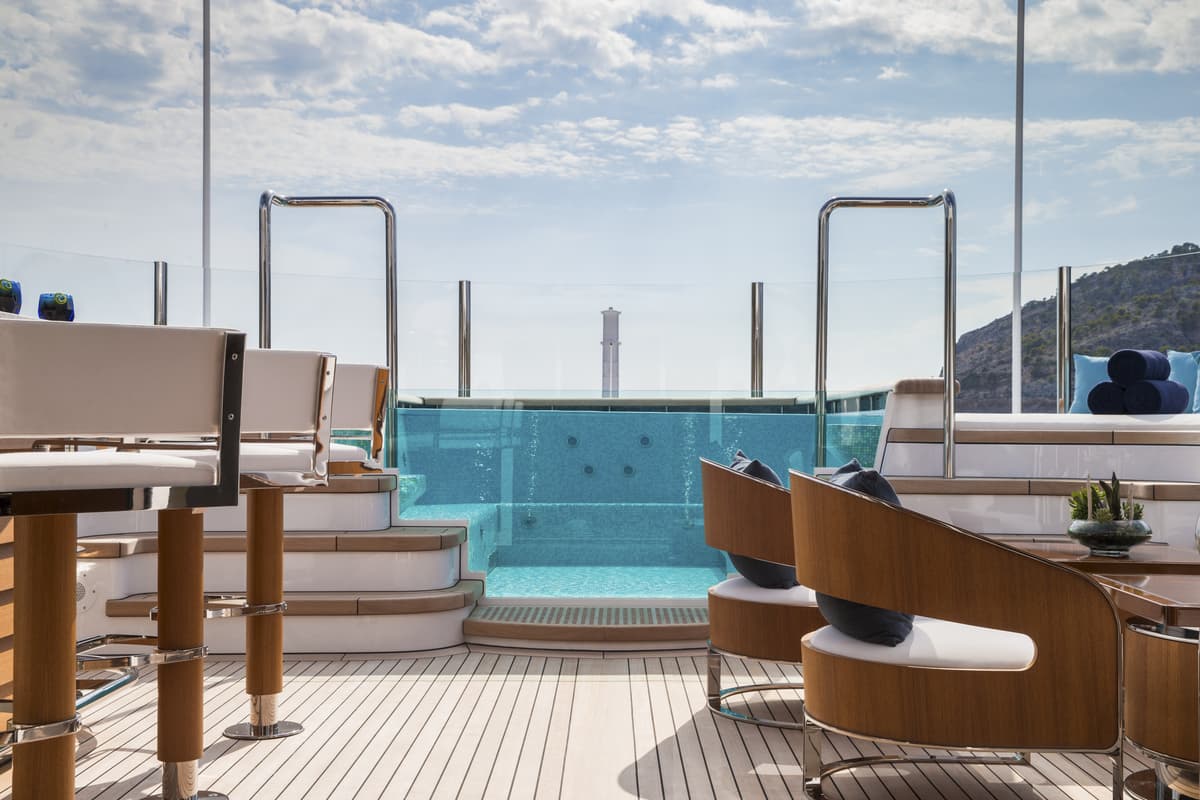 Aquila superyacht’s sun deck with a glass-fronted Jacuzzi, surrounded by cushioned seating and sun loungers, under a bright blue sky.