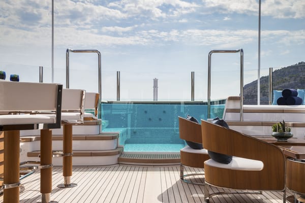 Aquila superyacht’s sun deck with a glass-fronted Jacuzzi, surrounded by cushioned seating and sun loungers, under a bright blue sky.