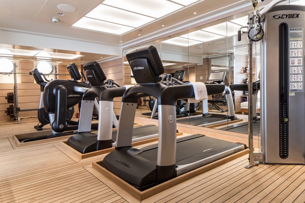 Aquila superyacht’s gym with treadmills, weight machines, mirrored walls, and light wood flooring under a bright, modern ceiling.