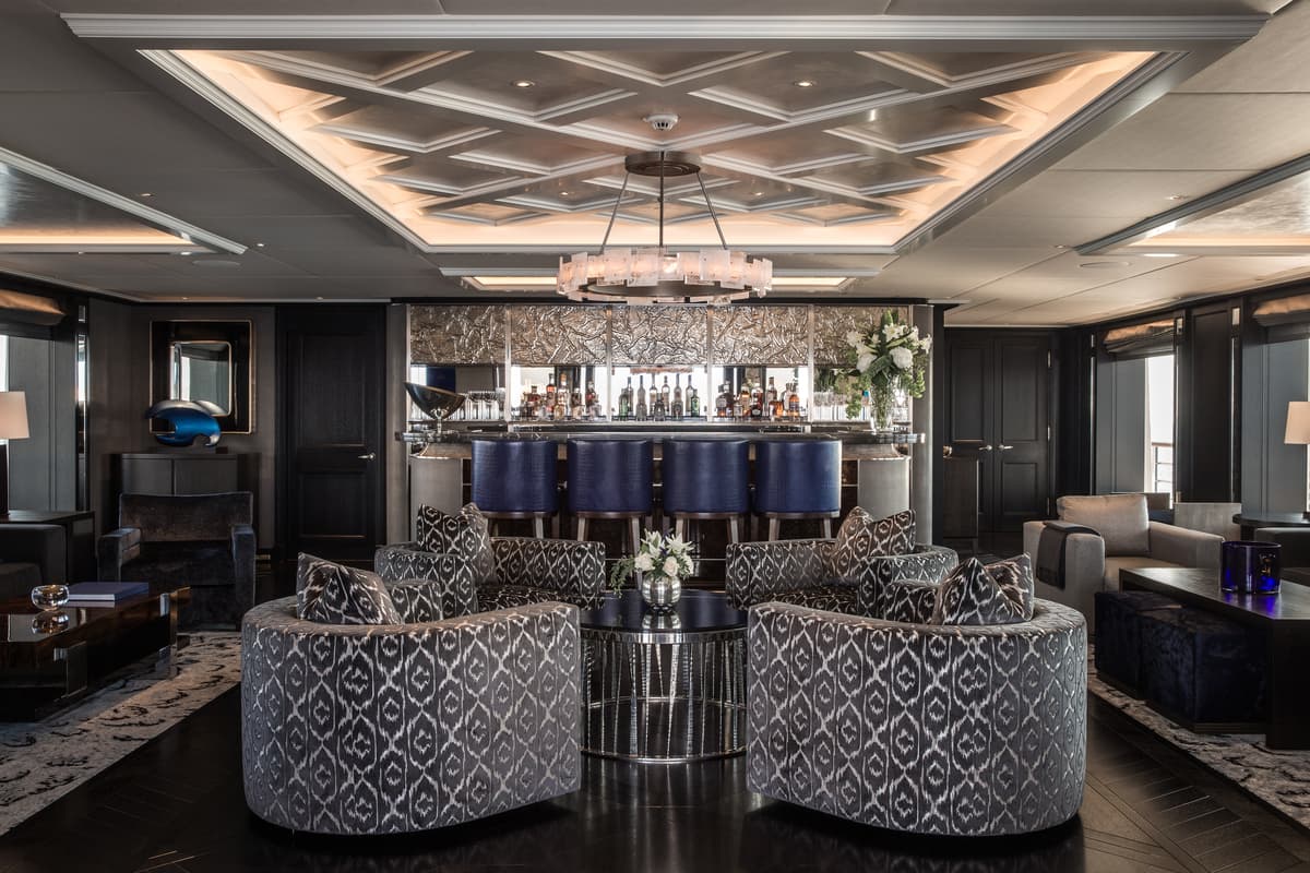 Aquila superyacht's luxurious owner’s saloon, featuring a central bar area with blue leather stools, patterned armchairs, and elegant modern decor.