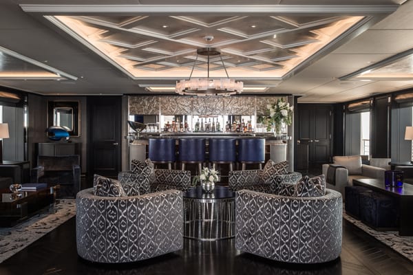 Aquila superyacht's luxurious owner’s saloon, featuring a central bar area with blue leather stools, patterned armchairs, and elegant modern decor.