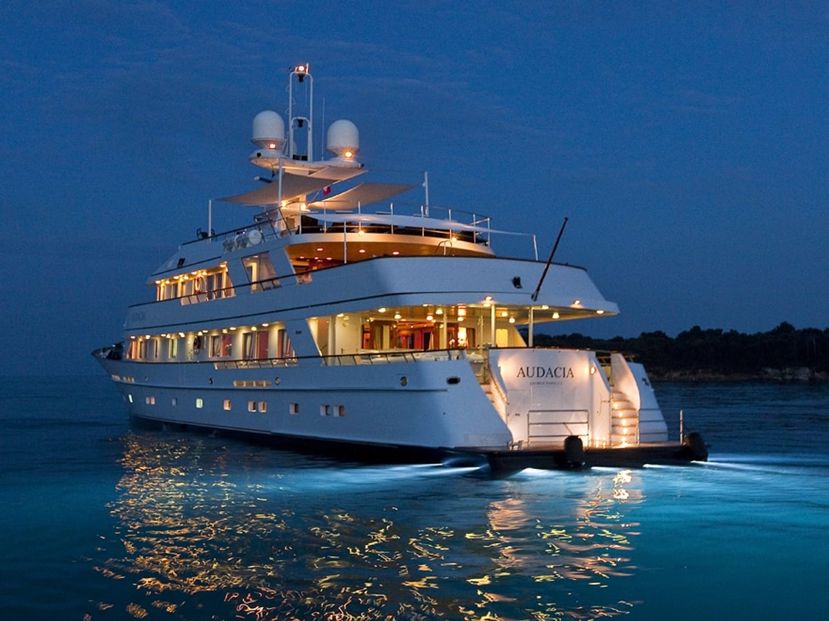 Audacia superyacht illuminated at dusk, with underwater lights reflecting on the calm water as she departs the coastline.