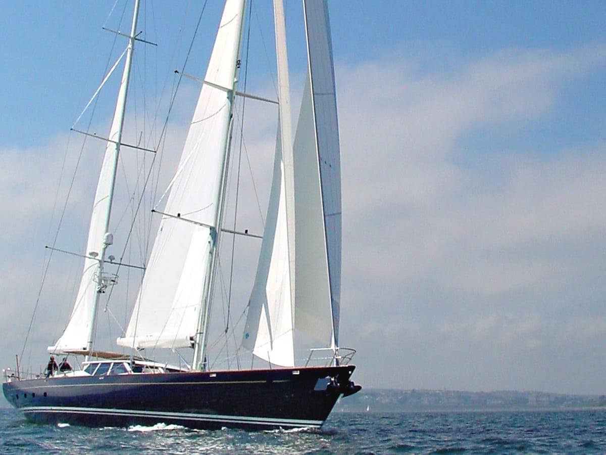 Beagle V sailing yacht under full sail on a clear day, gliding through calm blue waters with coastline in the distance.